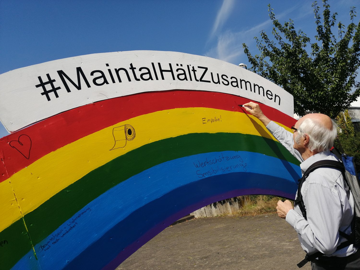Ein Passant beschriftet den farbenfrohen Regenbogen aus Holz, mit dem das Team der Maintal Aktiv – Freiwilligenagentur unter dem Motto #MaintalHältZusammen mit den Menschen ins Gespräch kommen möchte.
