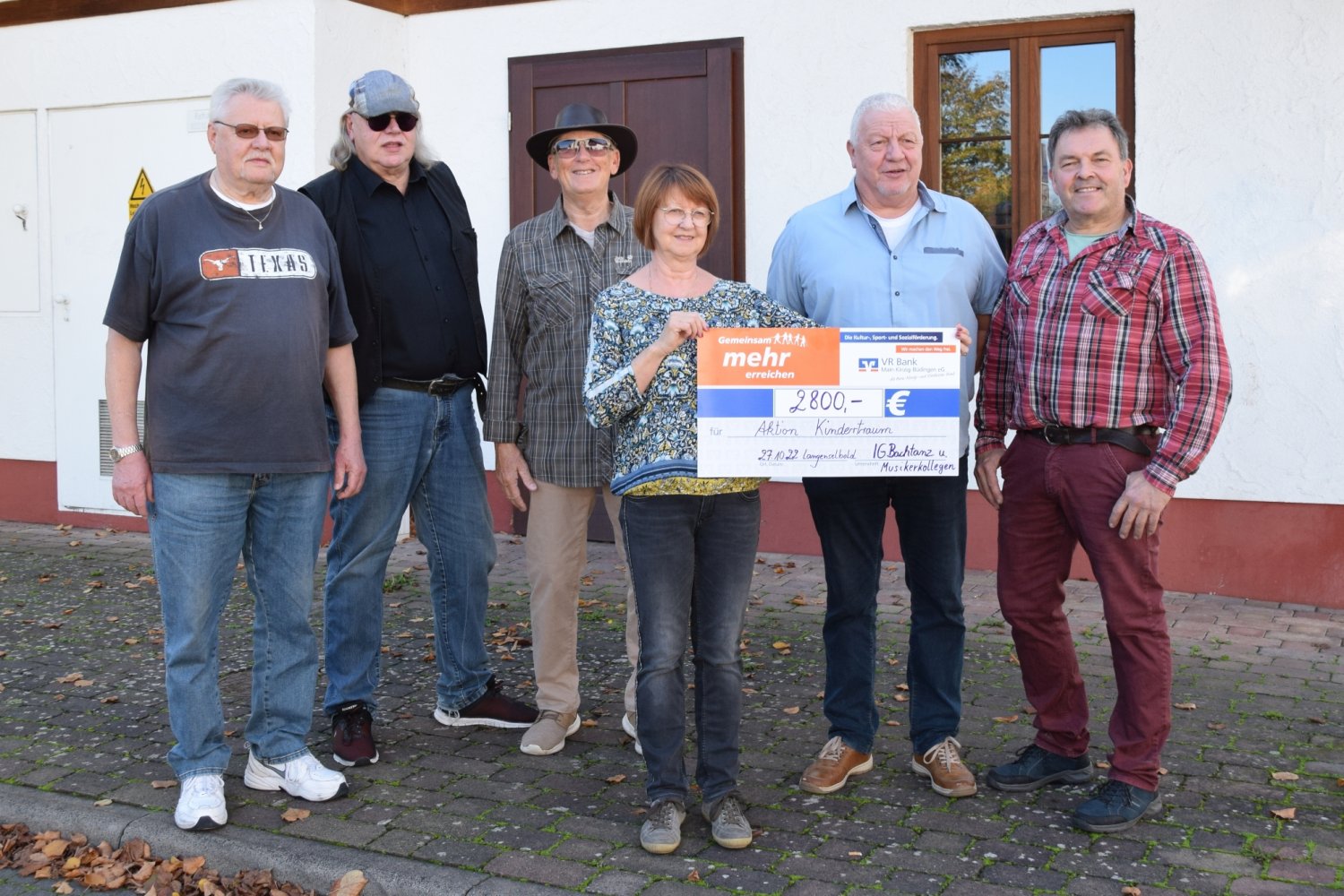 Lydia Semmel präsentiert vor dem Backhaus den Spendenscheck, eingerahmt von Mitgliedern der IG Bachtanz und den Musikern Volkmar Habermann (mit Cap) und Ian Browne (mit Hut)