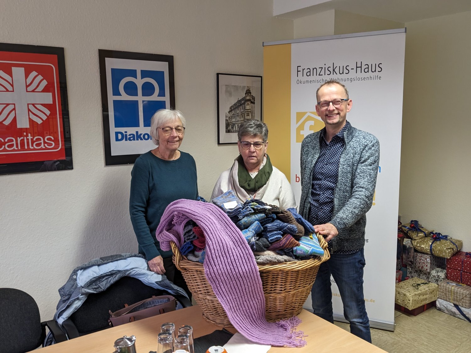 Cordula Stenger-Heynitz und Siegrid Kunkel von der Arbeiterwohlfahrt Bruchköbel überreichen einen ganzen Korb voller warmer Stricksachen an Einrichtungsleiter Rainer Broßmann vom Franziskus-Haus Hanau.