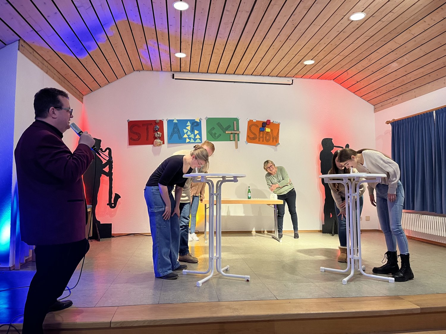 Die Kandidaten der Stapelshow beim Versuch, Zuckerwürfel mit dem Mund zu stapeln. Im Vordergrund: Moderator Michael Weigand, im Hintergrund sichtlich amüsiert: Brigitte Wolf. 