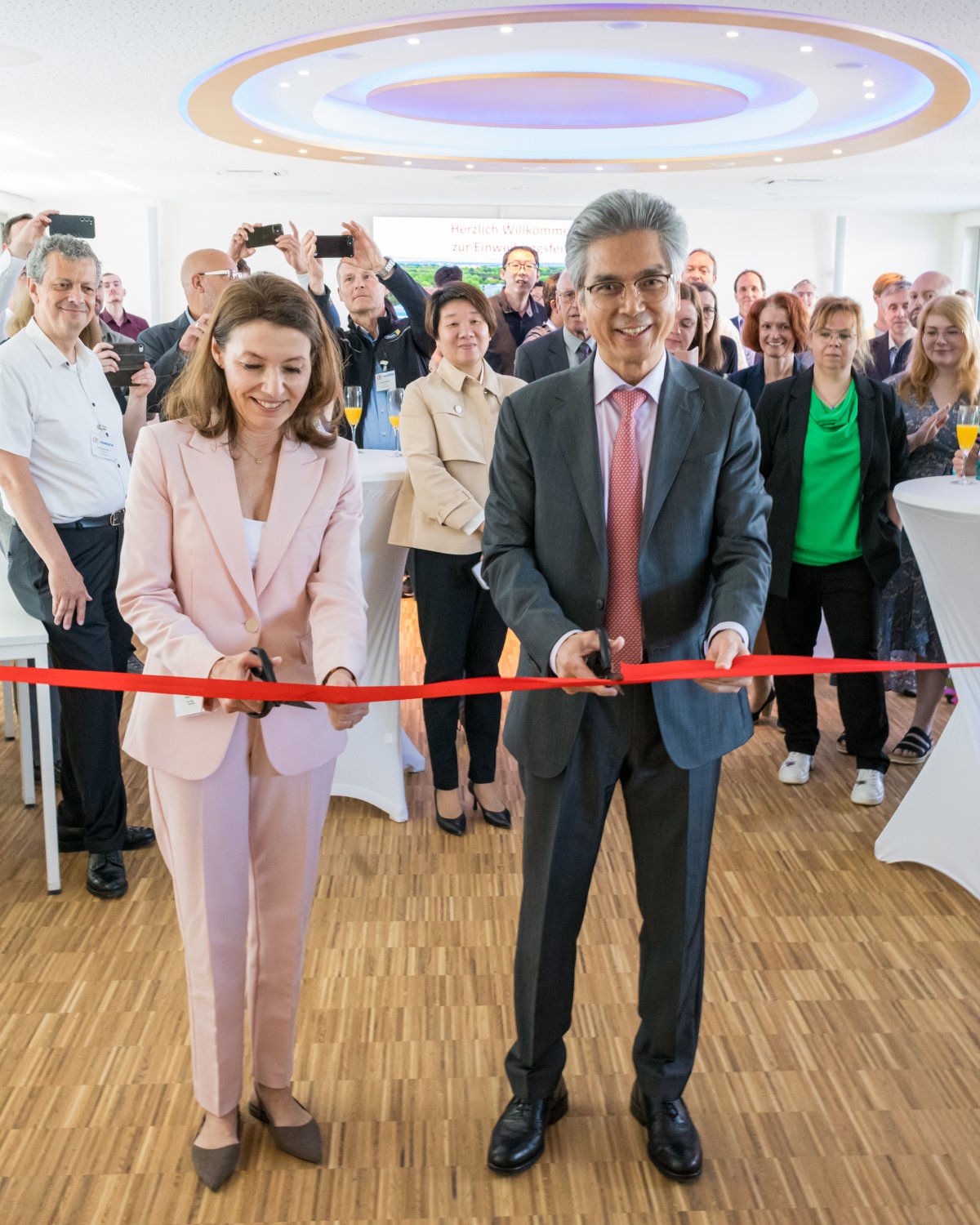 Jiaqiang Chen (Geschäftsführer Hongfa Europe GmbH) und Bürgermeisterin Monika Böttcher zerschneiden gemeinsam das rote Band und weihen damit das neue Vertriebsgebäude der Hongfa Europe-Zentrale am Marie-Curie-Ring ein.